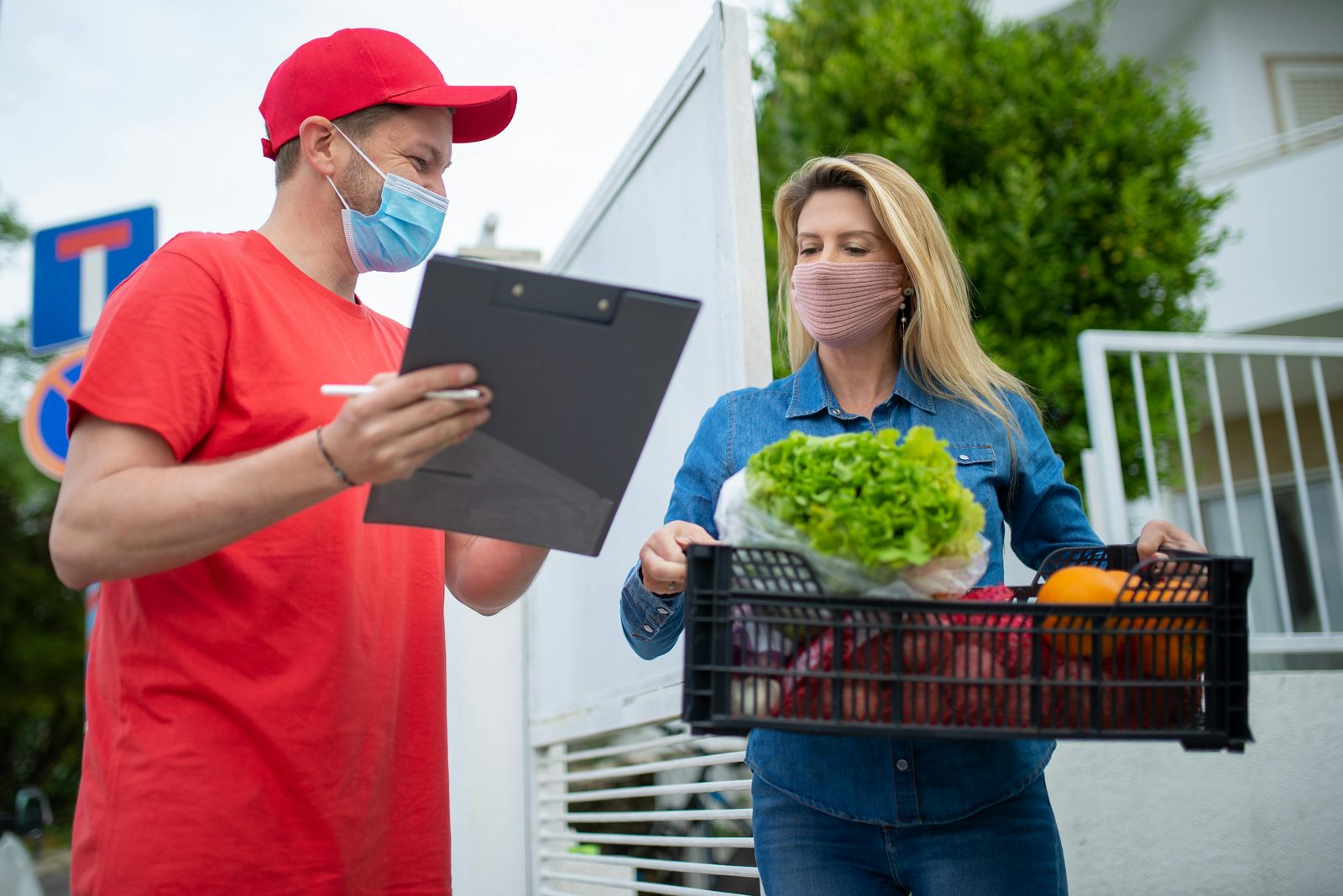 A delivery driver with a clipboard handing a crate of groceries to a customer at her gate