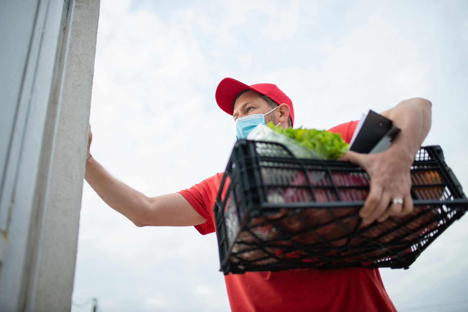 A delivery driver carrying a crate of groceries and a clipboard to a customer's door