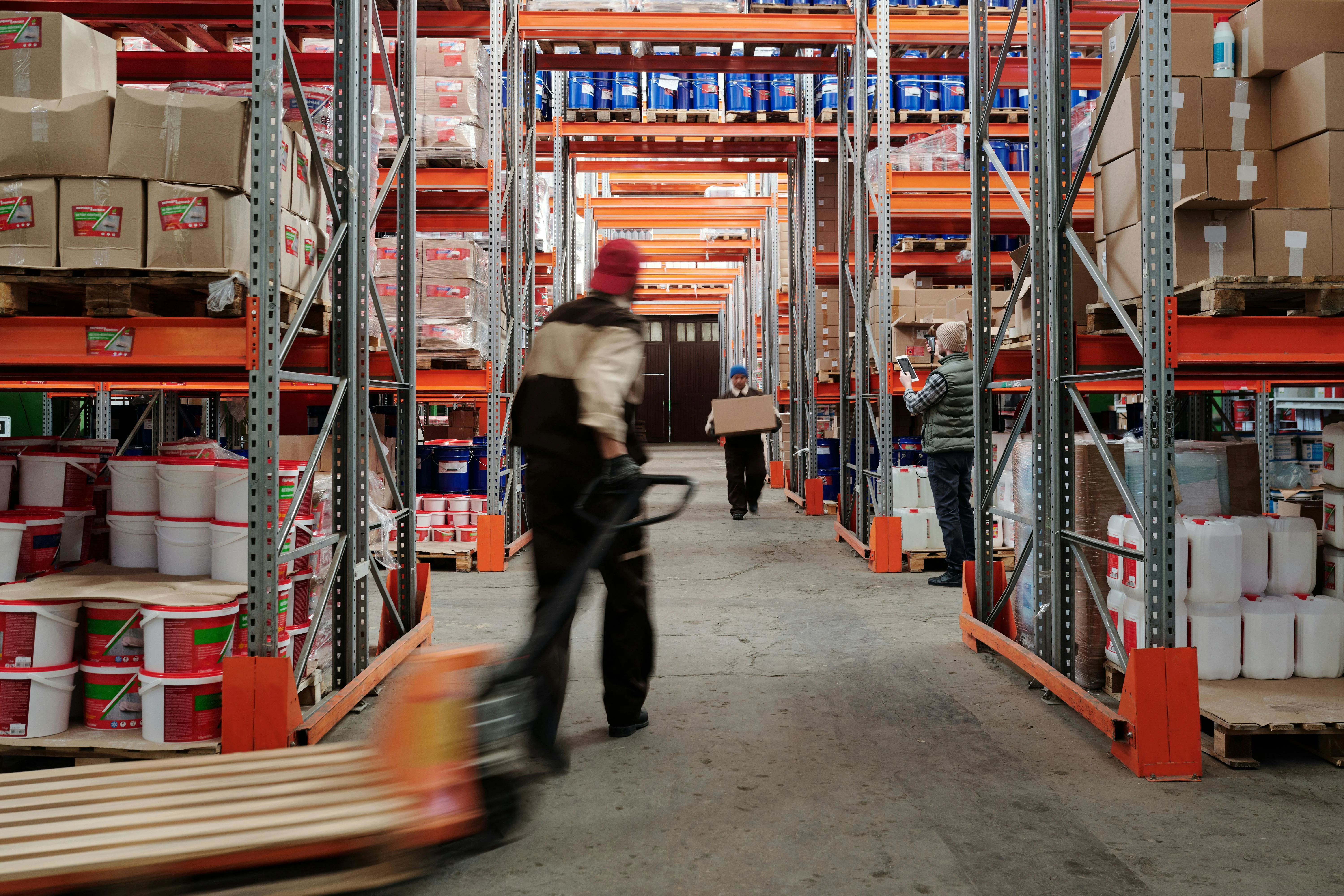 Workers moving stock along a warehouse aisle, one pulling a pallet jack