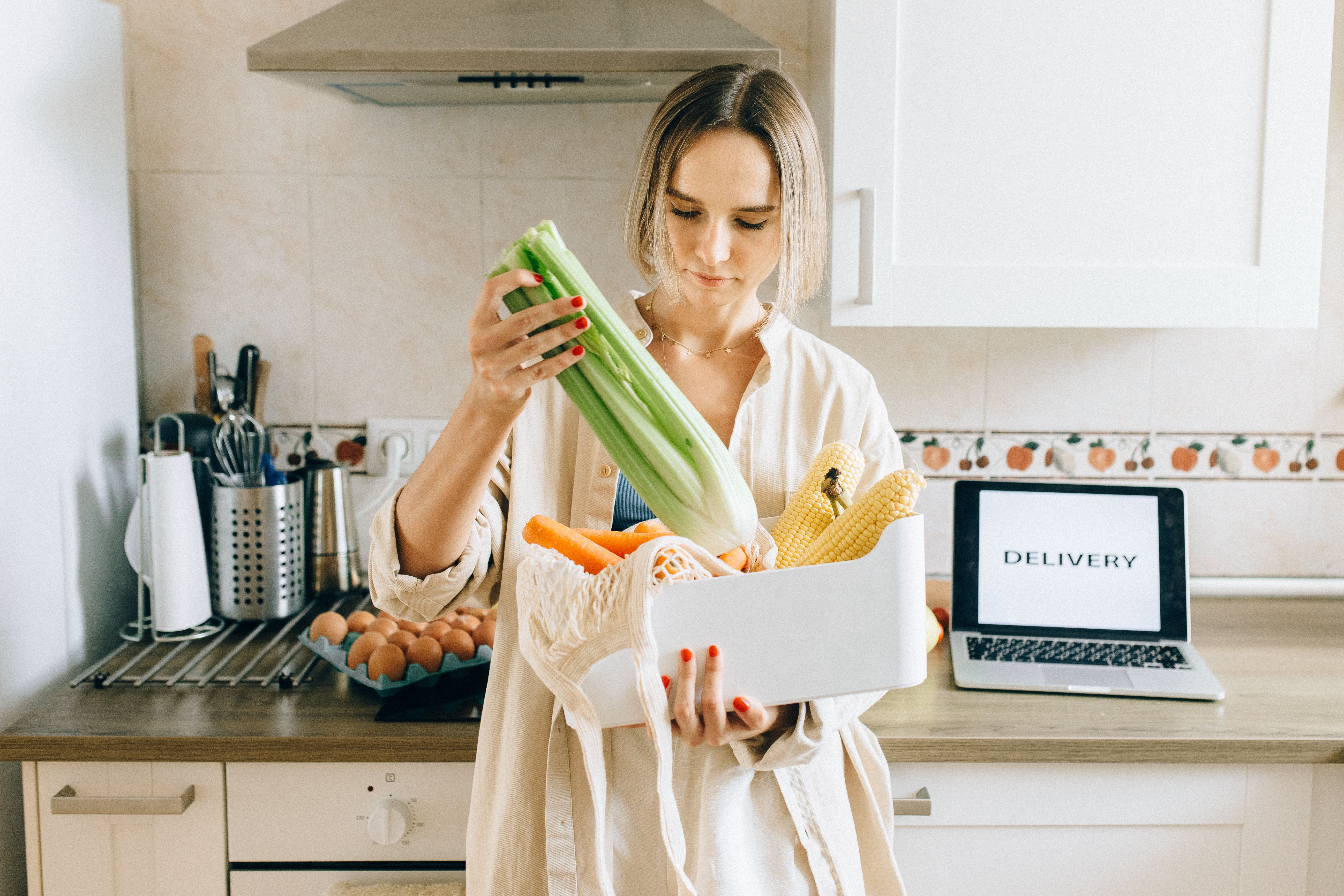 A woman unpacks celery, carrots and corn from a delivery box in her kitchen, with a laptop showing the word Delivery.