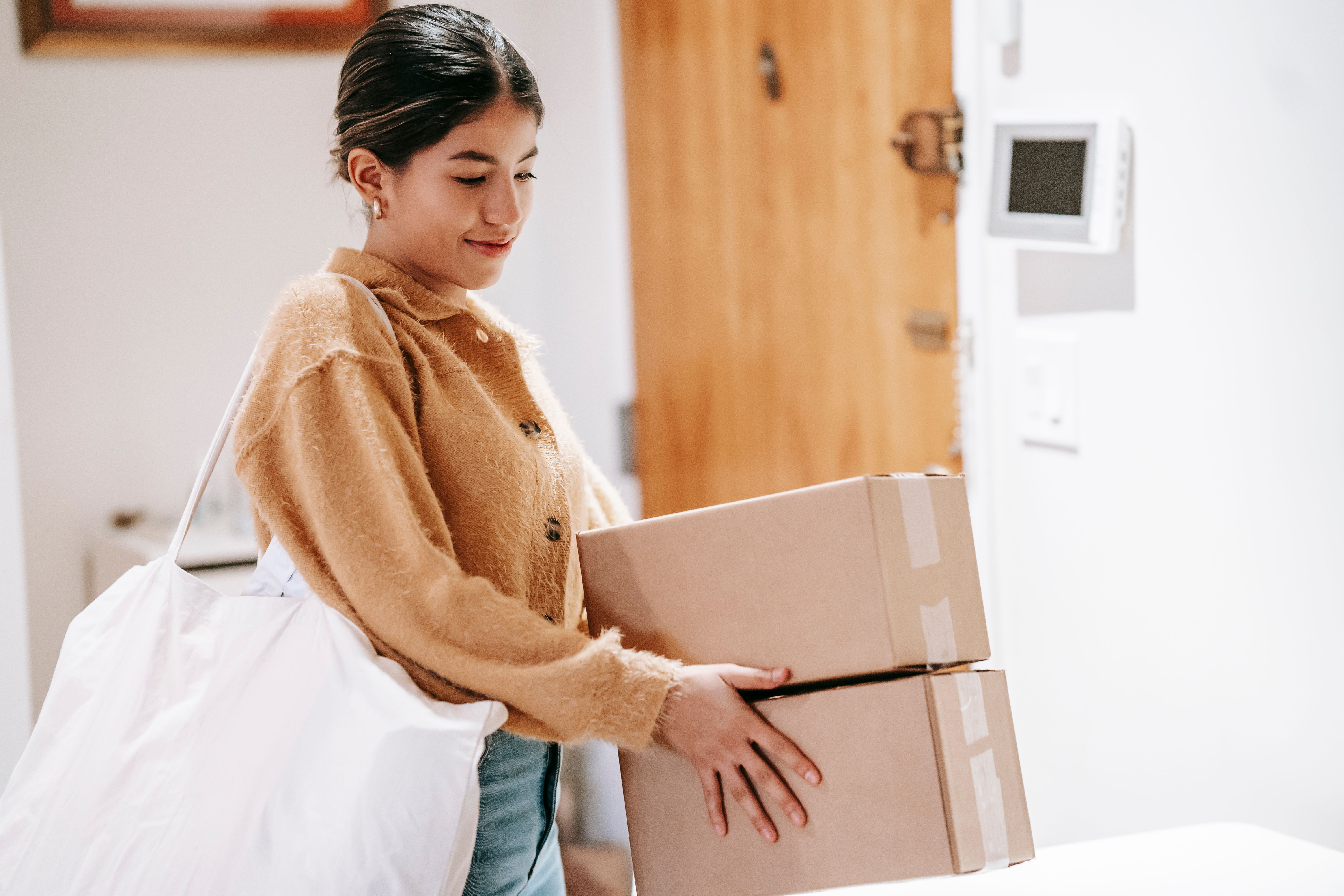 A smiling woman carries two cardboard parcels into her home.