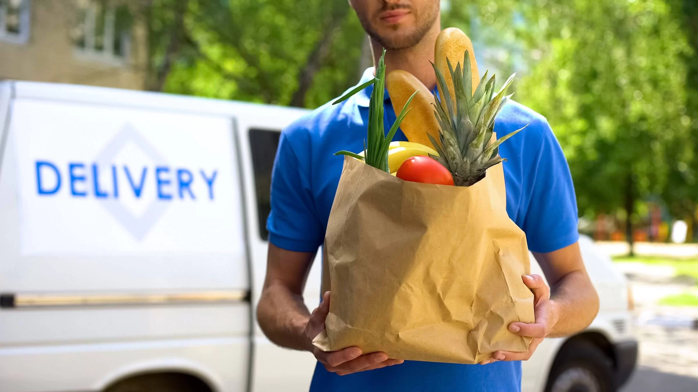 A delivery driver holding a paper bag of groceries next to a delivery van