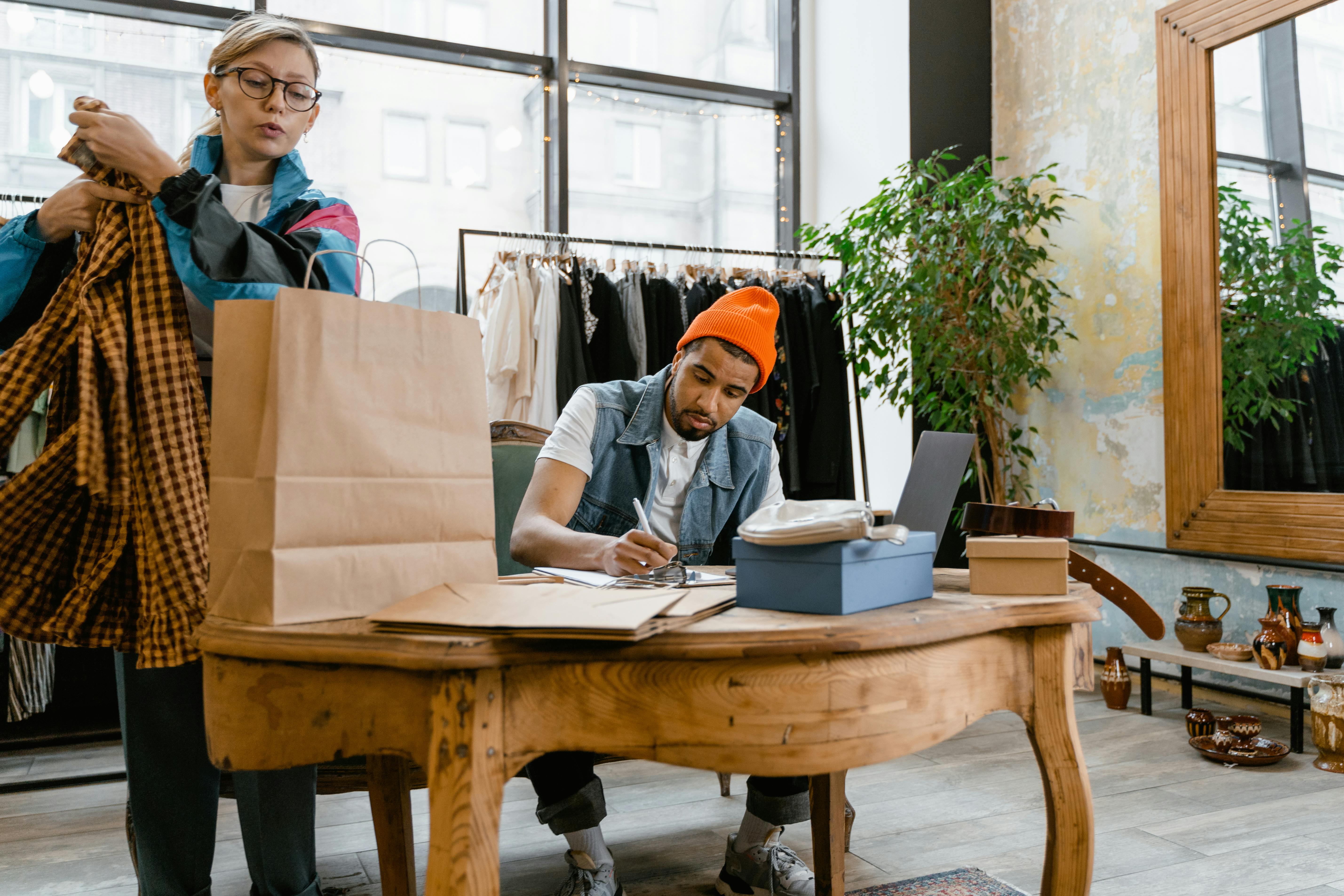 Two staff in a clothing boutique, one packing an order into a paper bag while the other writes notes next to a laptop