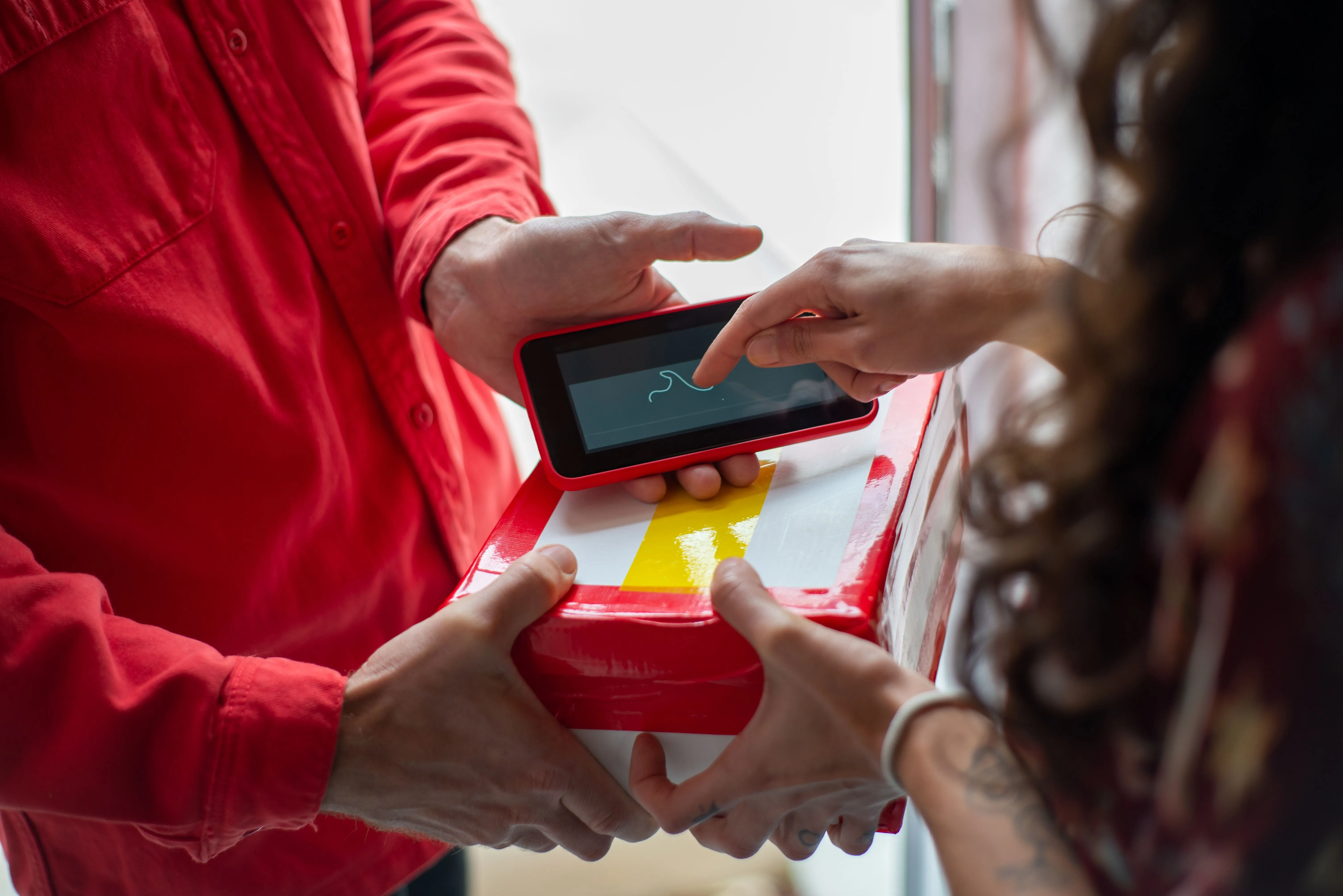 A delivery person in a red shirt holds out a card reader while a customer taps it over a red delivery box.