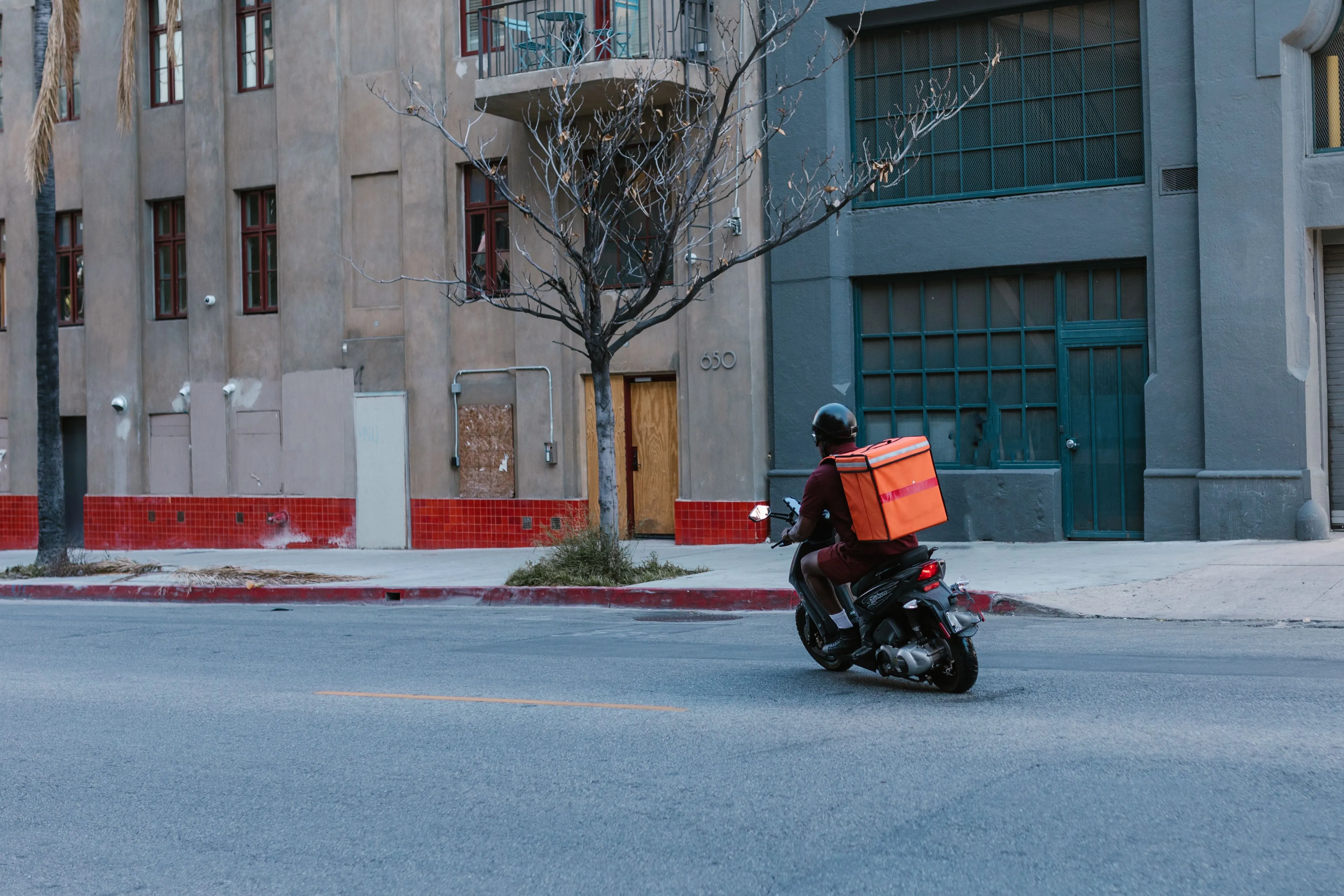 A delivery rider with an orange insulated box rides a scooter down a city street.
