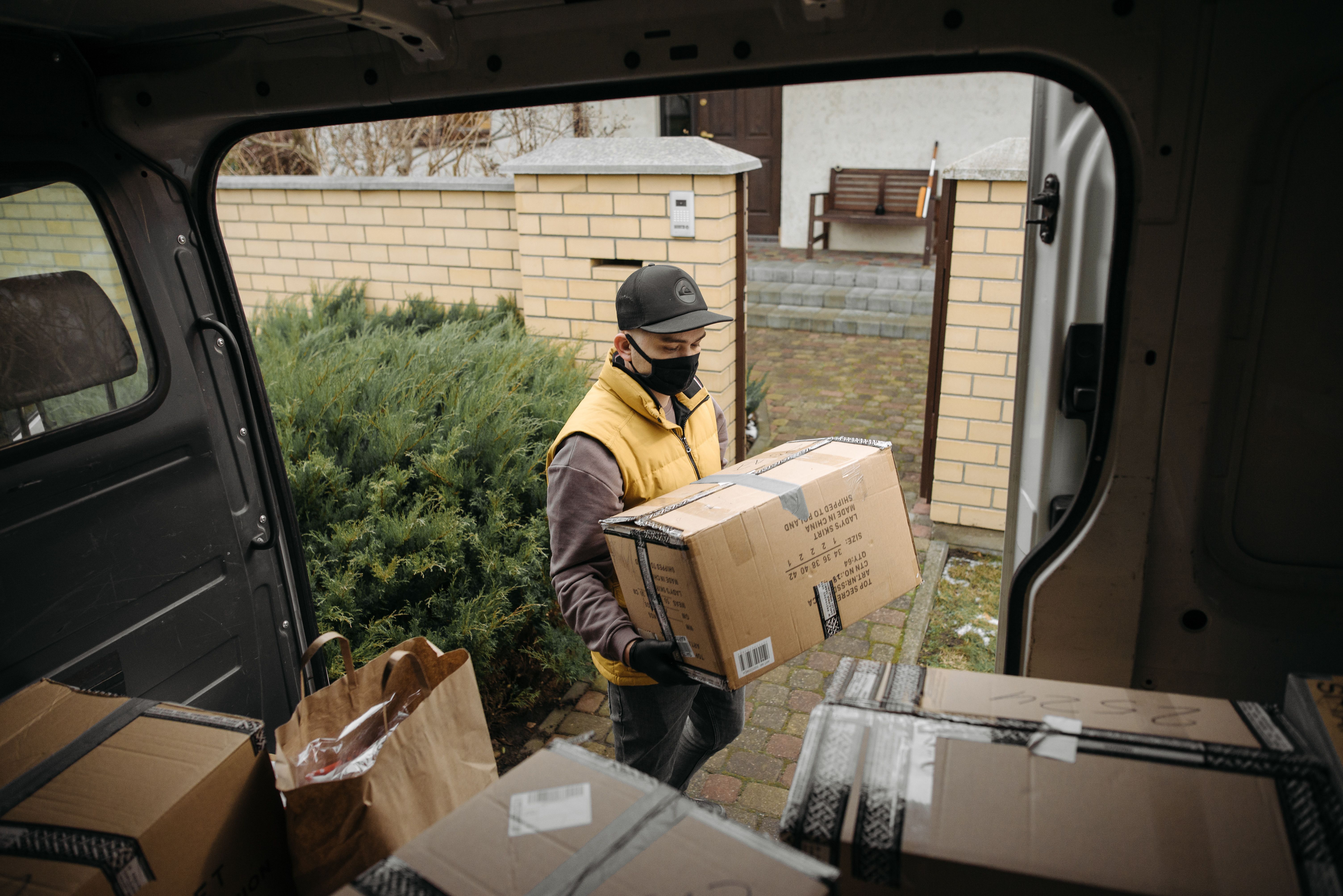 A courier in a face mask lifts a cardboard box from a van loaded with parcels outside a house.