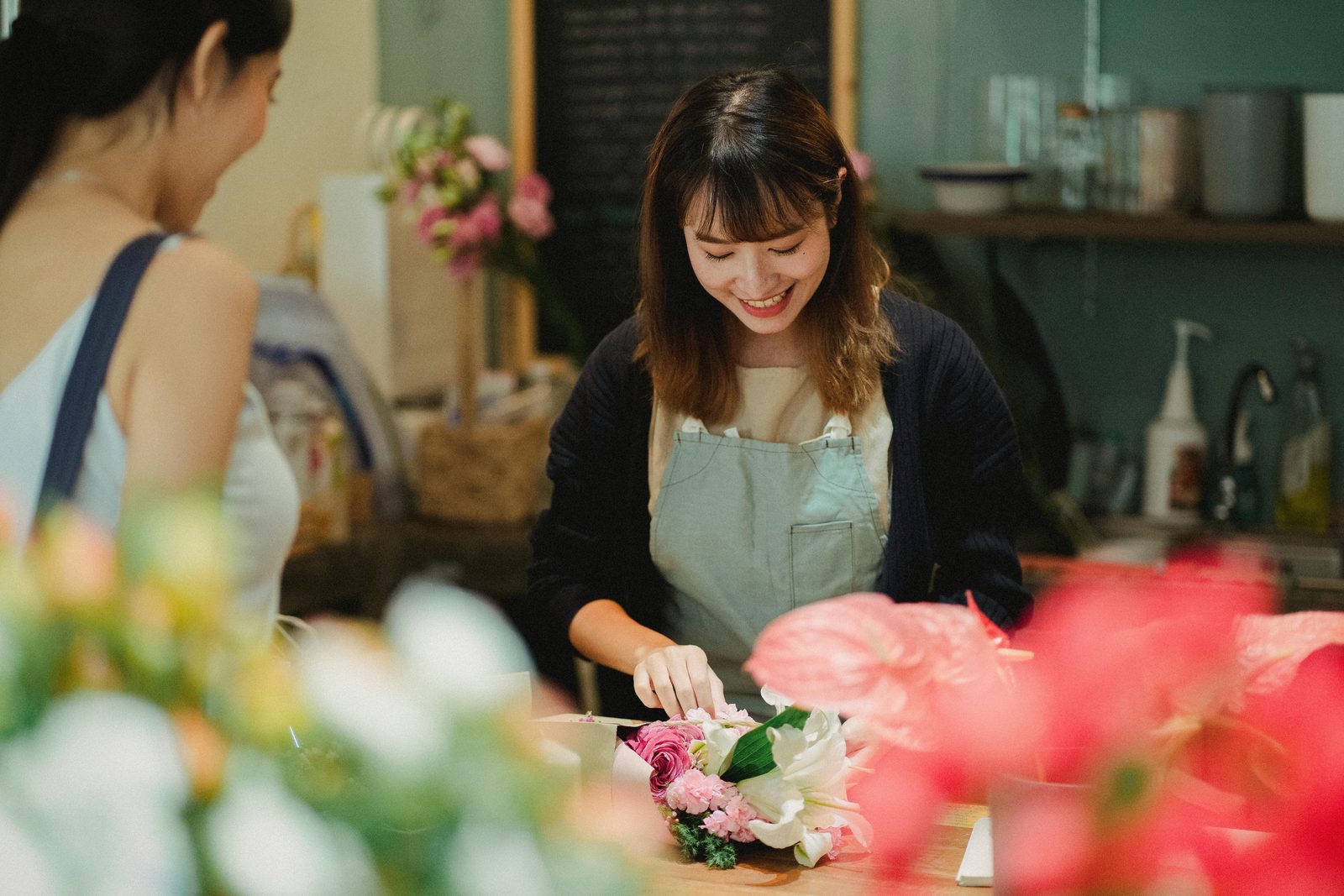 A florist wrapping a bouquet of pink roses and white lilies at the shop counter while a customer waits