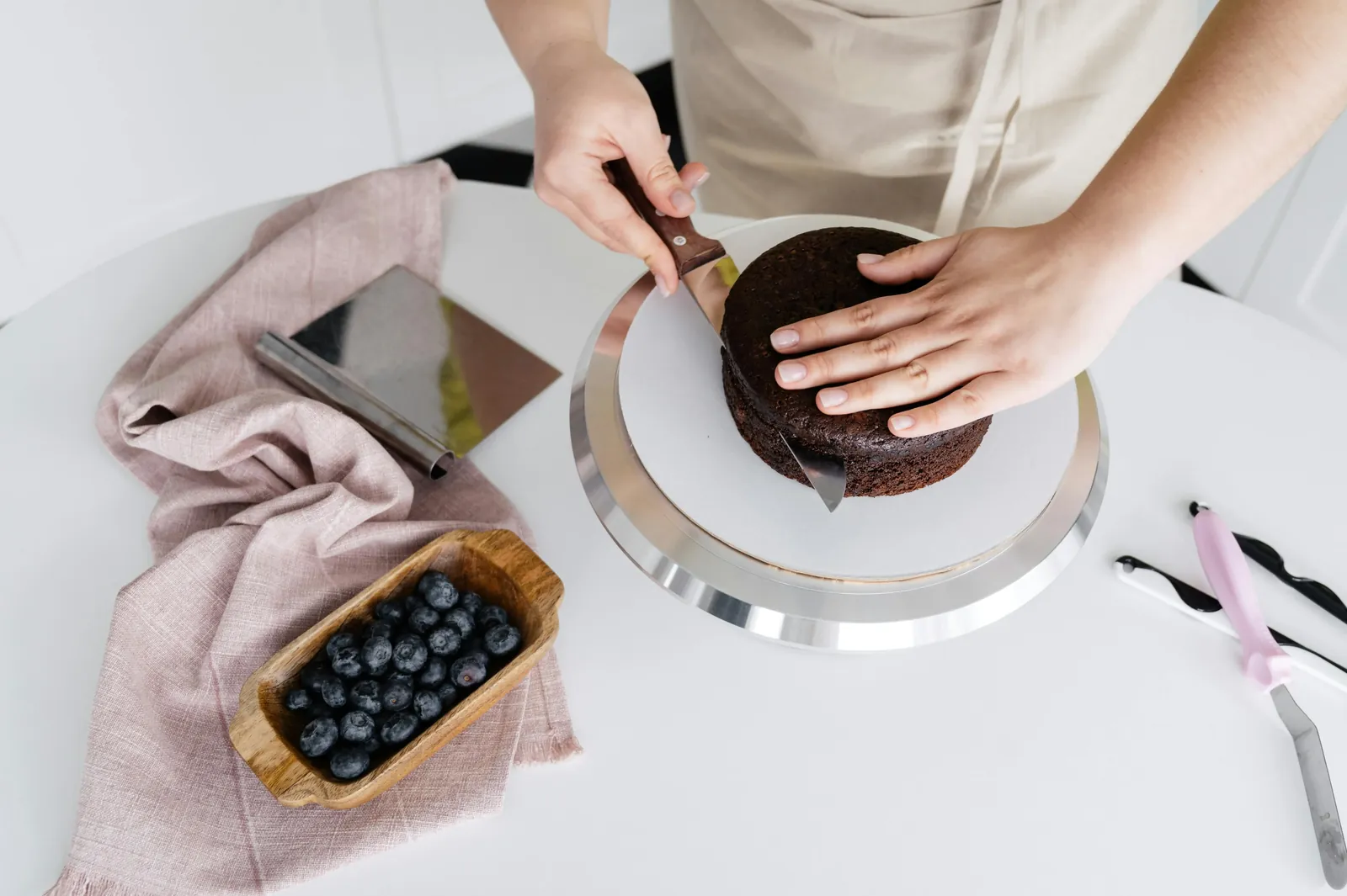 A baker trimming a chocolate sponge cake on a turntable, with a bowl of blueberries beside it