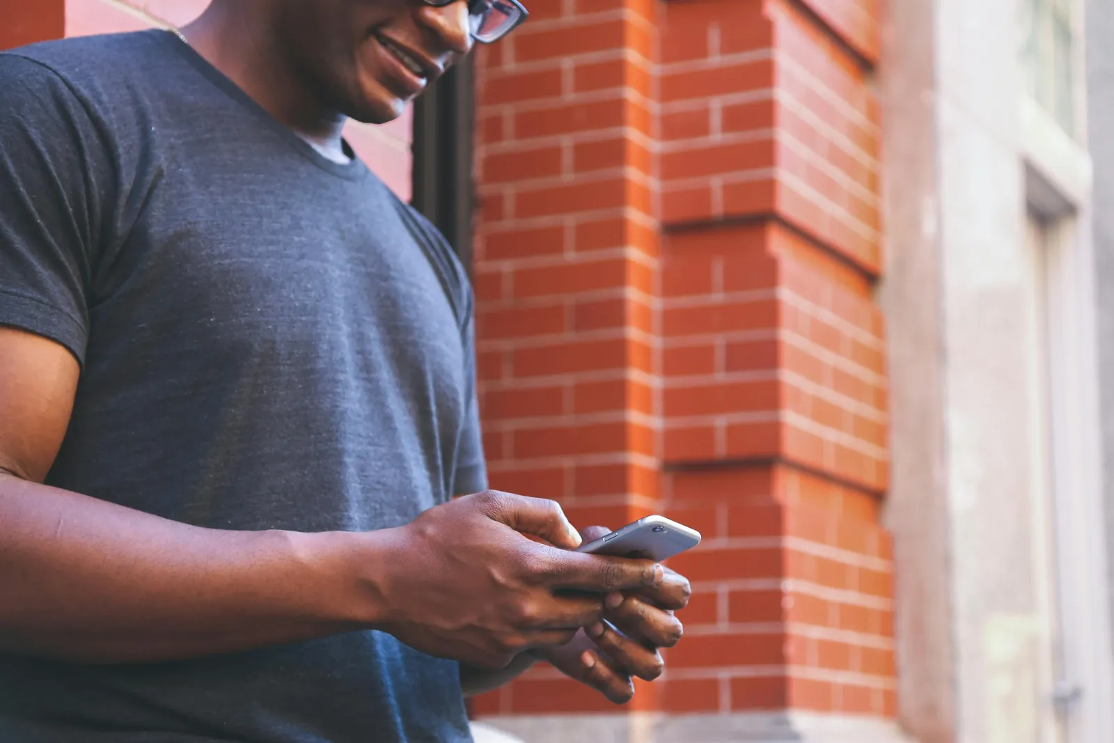 A smiling man in a T-shirt checks his phone outside a brick building