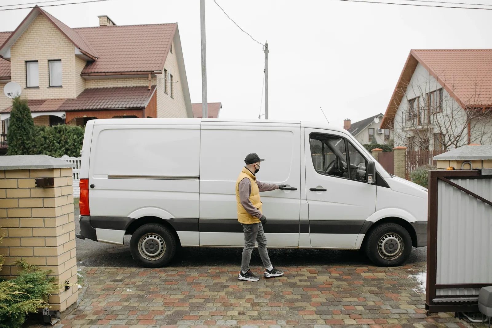 A delivery driver in a yellow vest walks to the side door of a white van parked in a residential driveway