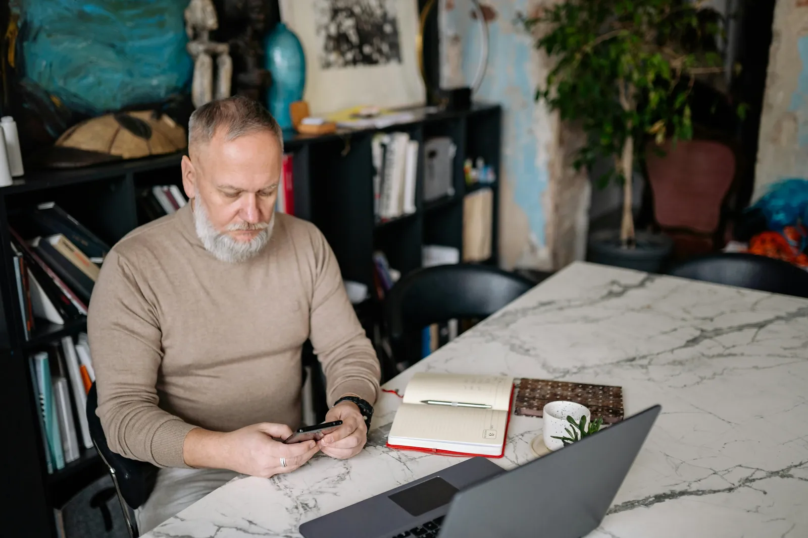 A man at a marble table checks his phone, with a laptop and an open notebook in front of him