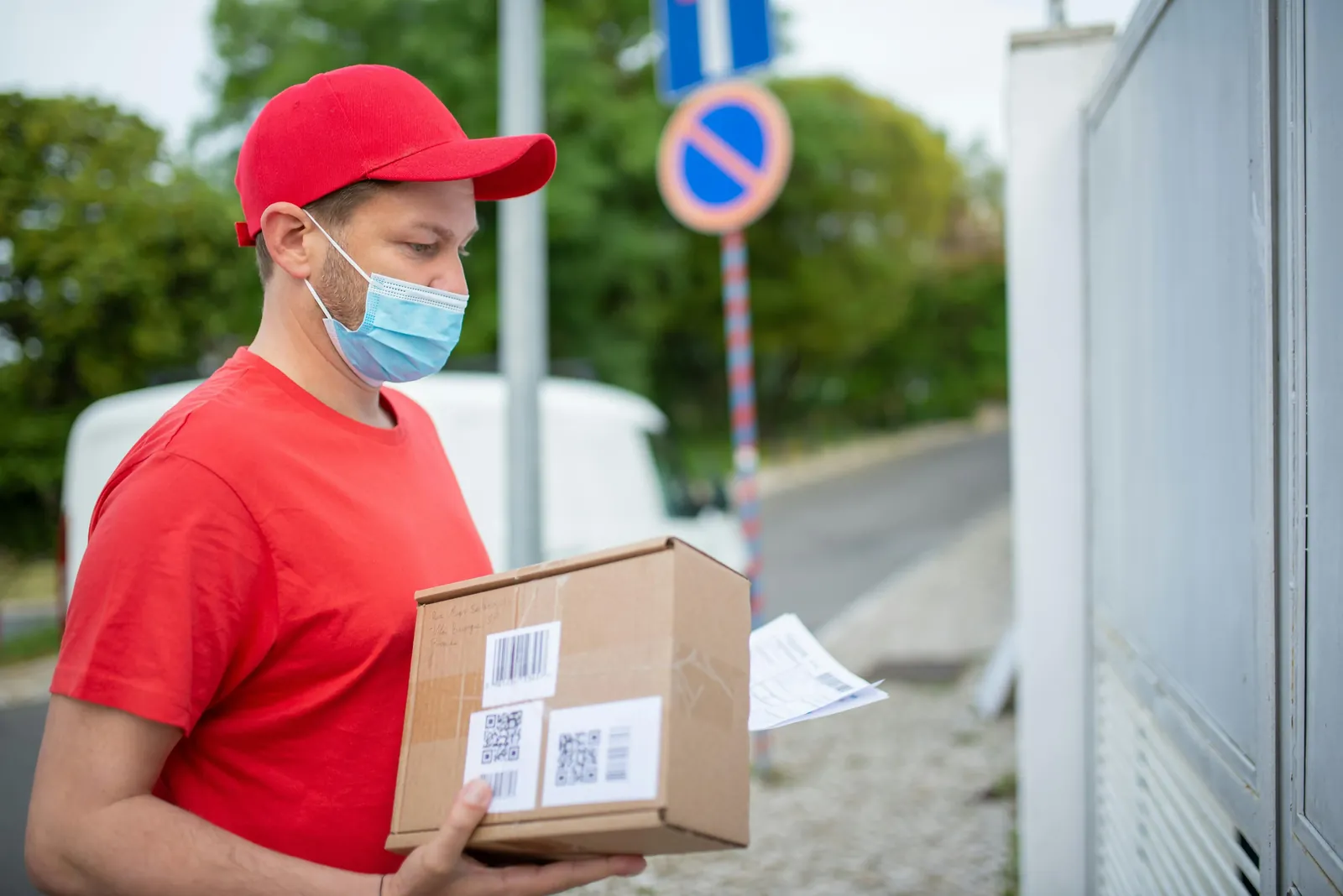 A delivery driver in a red cap and face mask checks a parcel's label outside a gate.