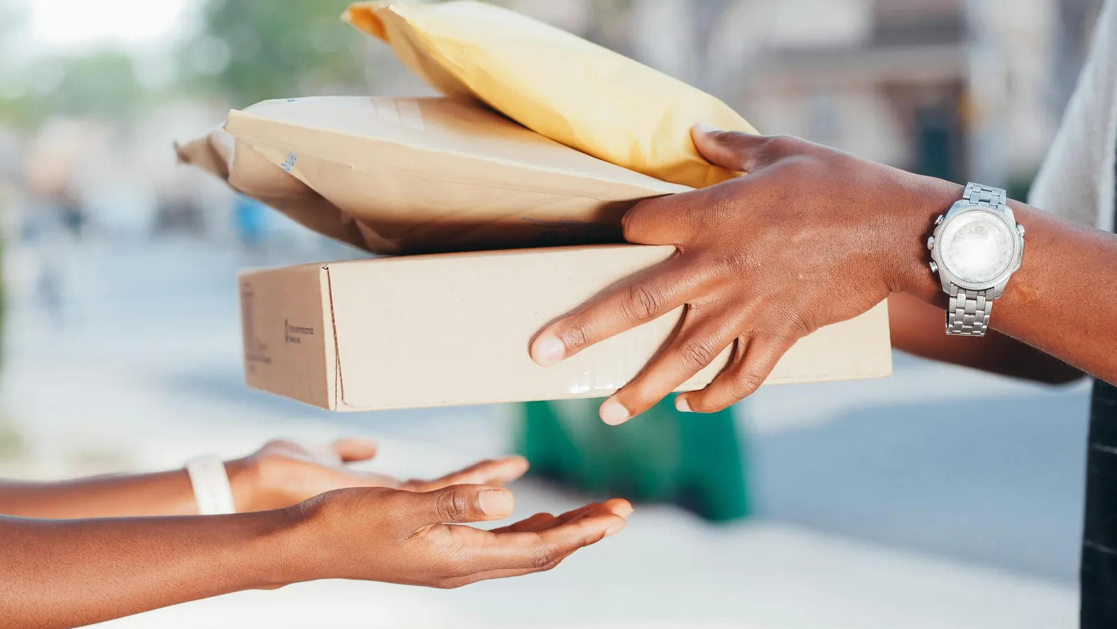 A courier's hands pass a cardboard box and two padded mailers into a customer's open hands on a sunny street.