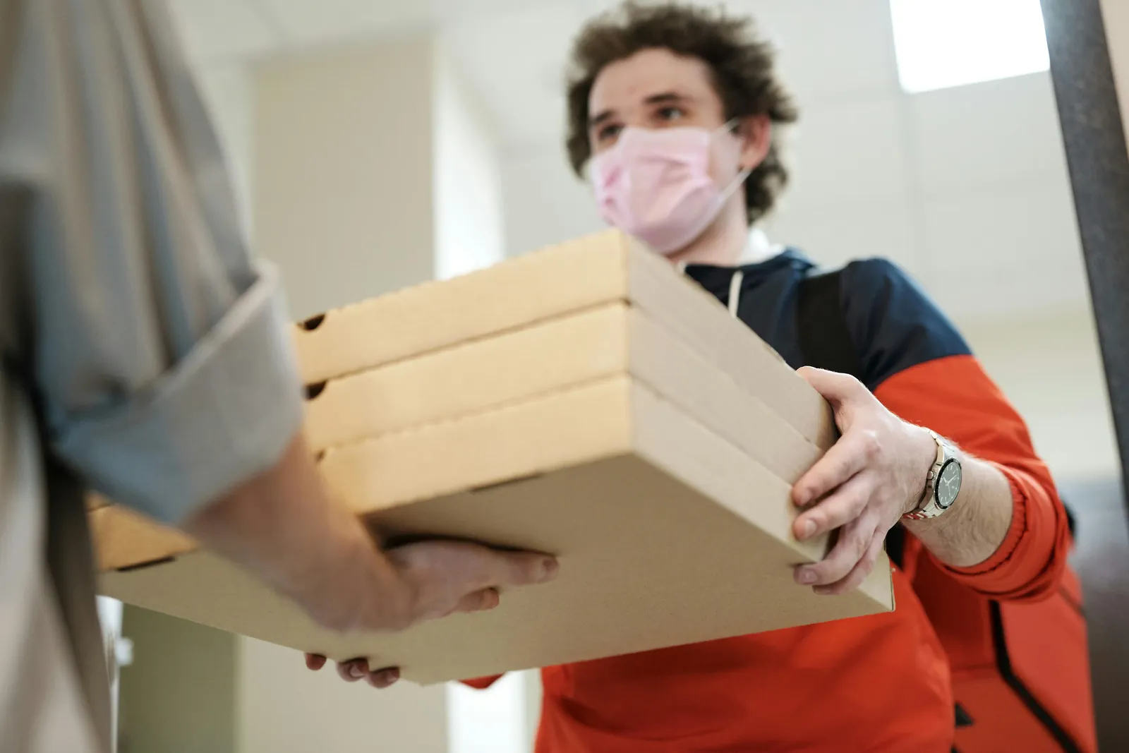 A courier in a face mask hands a stack of pizza boxes to a customer at the door.