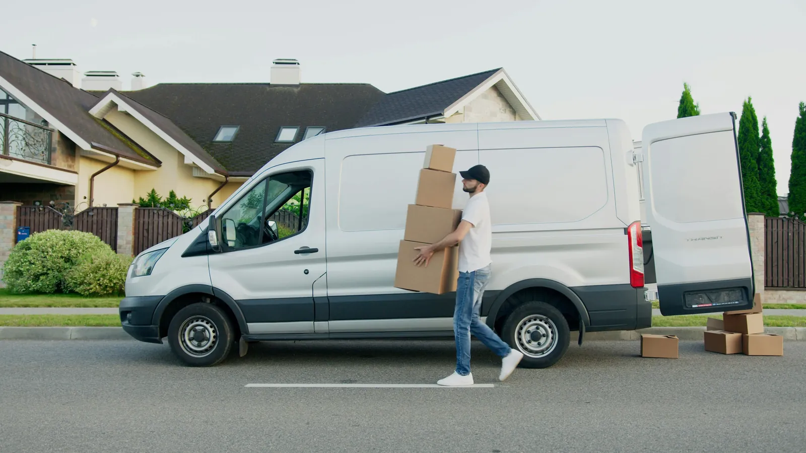 A delivery driver carries a stack of cardboard boxes past a white van parked outside a house